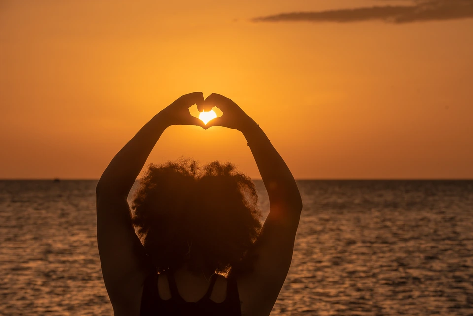 Silhouette forming a heart with raised hands against a sunset