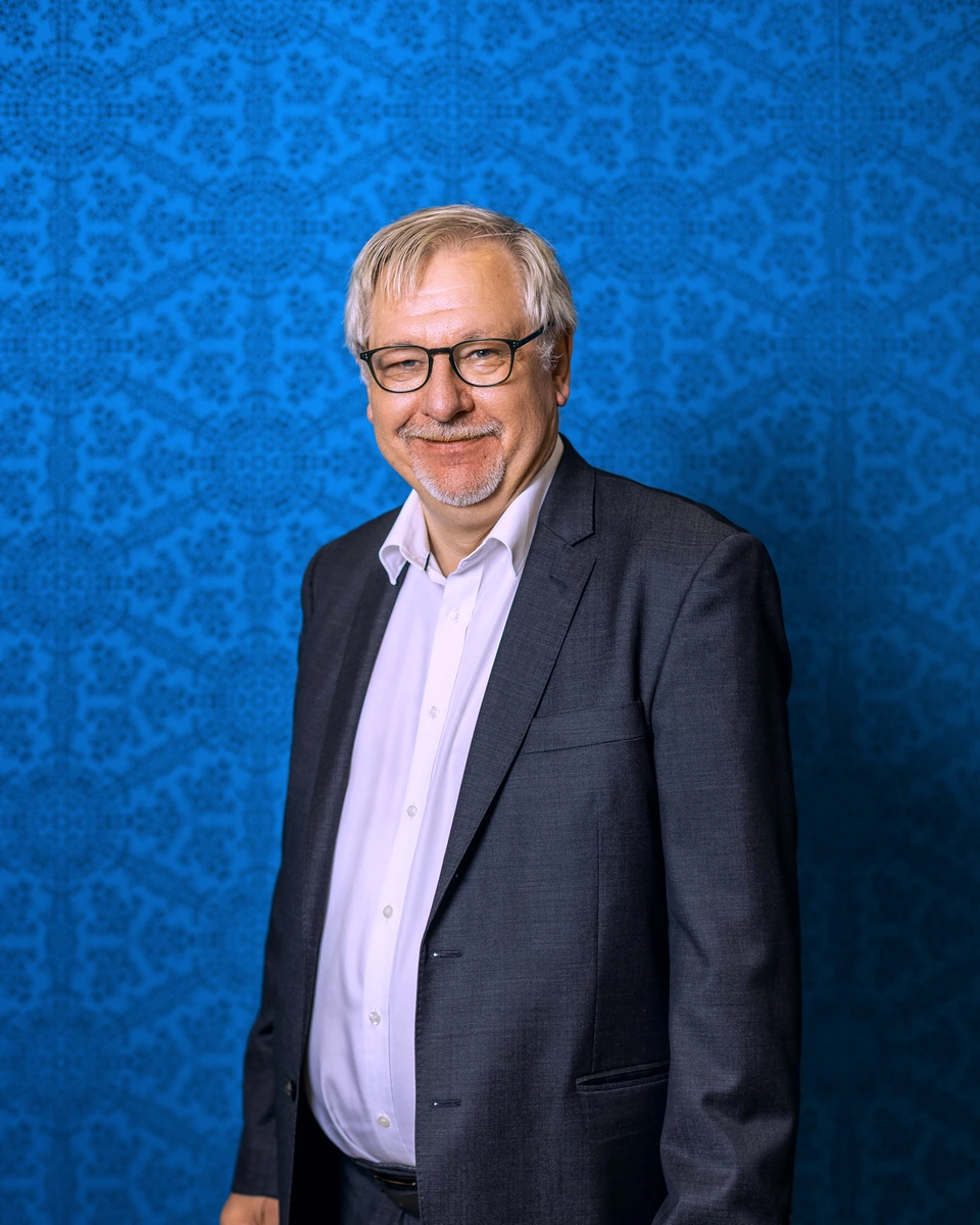 Man wearing a dark jacket photographed against a blue patterned wall