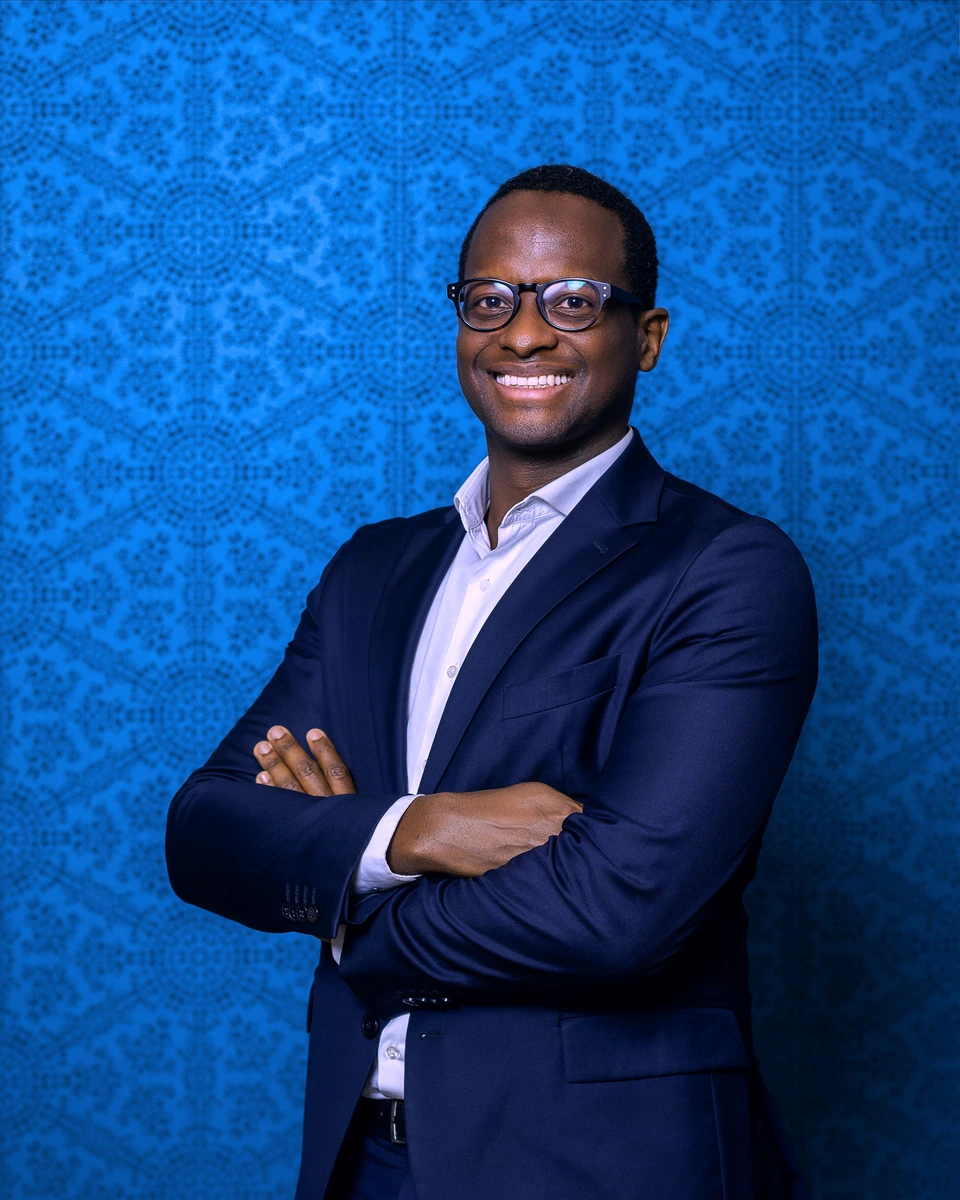 Man in a navy suit standing with folded arms against a blue backdrop